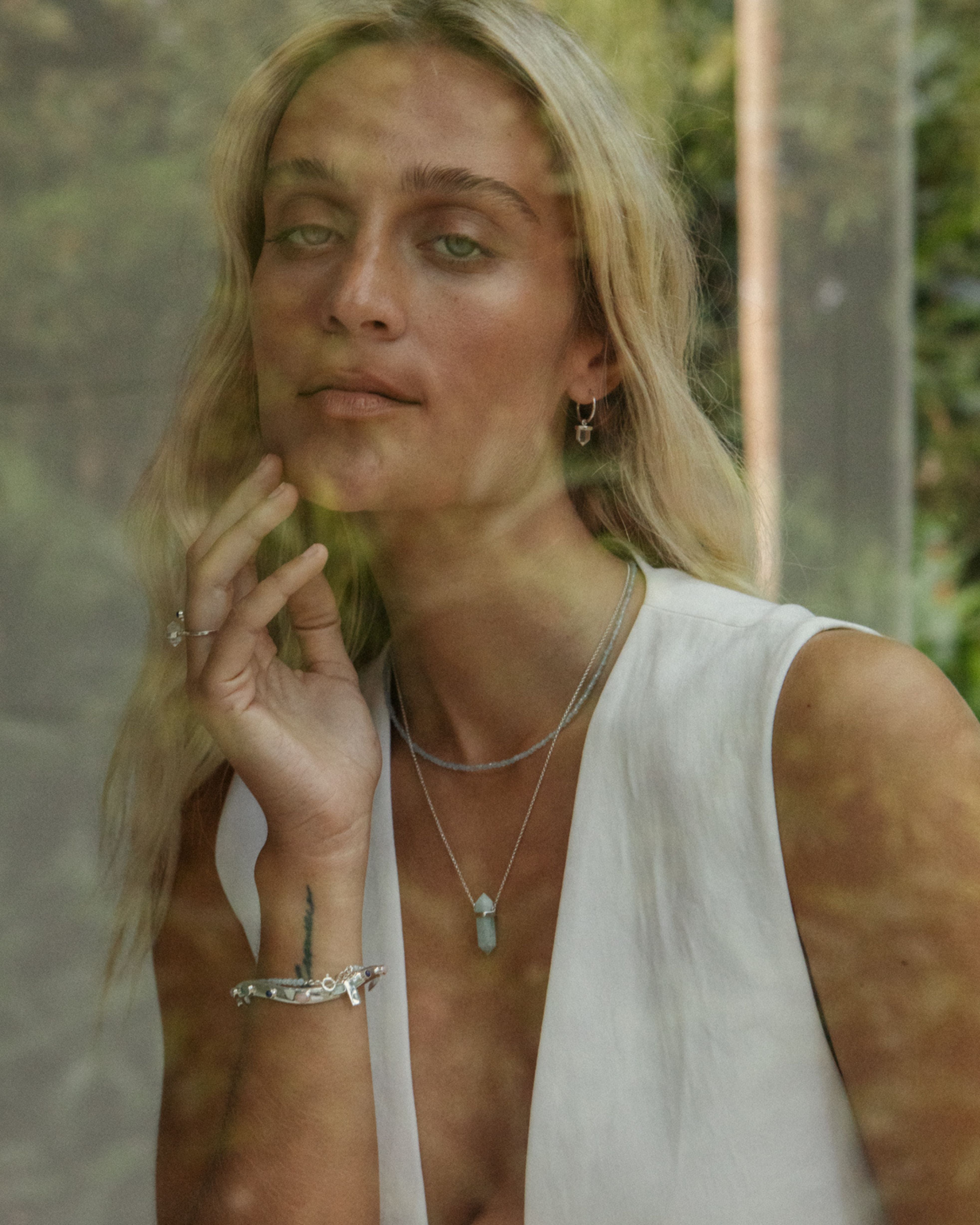 Woman with blonde hair and a white sleeveless top, surrounded by greenery.