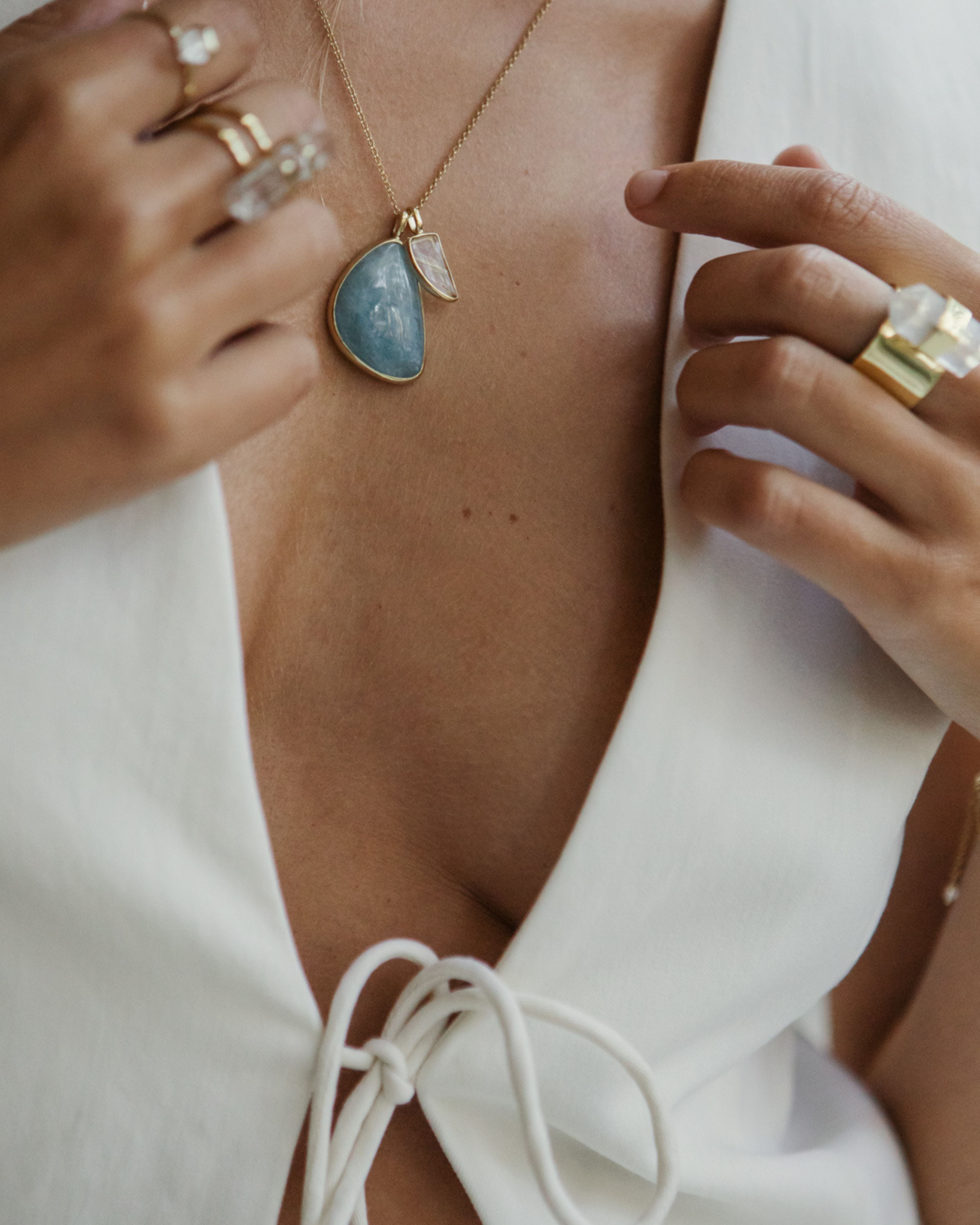Close-up of a person wearing a necklace with a Larimar + Rutilated Quartz and gold rings on a white background